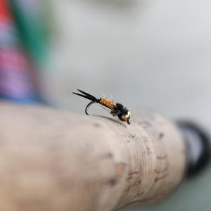 May include: A close-up of a black and orange fly fishing lure with a gold bead on a cork fishing rod.