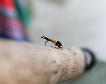 May include: A close-up of a black and orange fly fishing lure with a gold bead on a cork fishing rod.