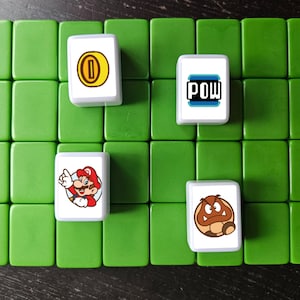 May include: A set of six white tiles with different images from the Super Mario Bros. video game franchise. The tiles feature a gold coin, a red mushroom, a green turtle shell, a character of Mario, and the word "POW!" in a blue and white box.