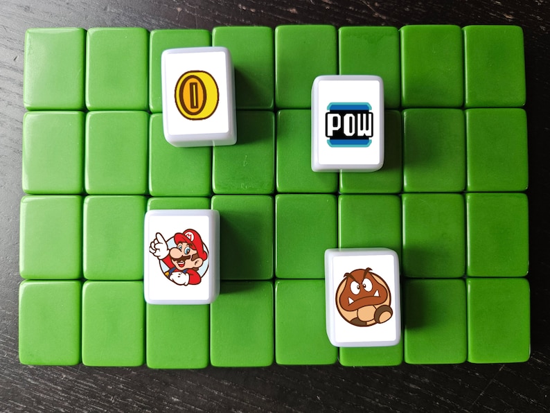 May include: A set of six white tiles with different images from the Super Mario Bros. video game franchise. The tiles feature a gold coin, a red mushroom, a green turtle shell, a character of Mario, and the word "POW!" in a blue and white box.