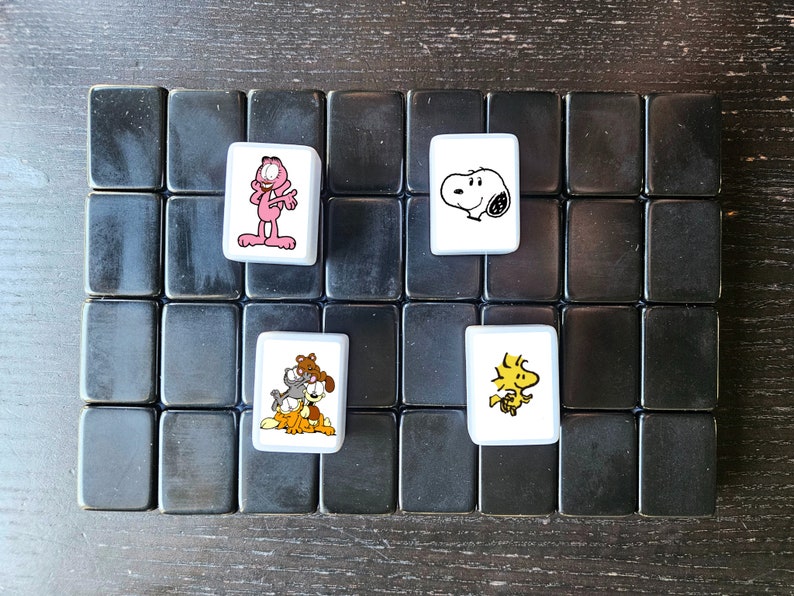 May include: A black and white game board with four white tiles. Each tile features a cartoon character. One tile has a pink cat, one has a black and white dog, one has a yellow bird, and one has a white dog.