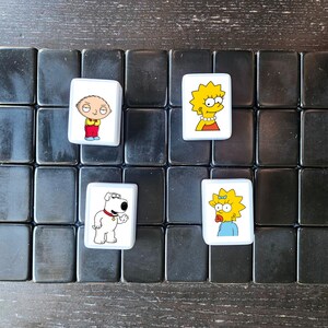 以下が含まれることがあります： 白黒のゲームボードに白いタイルが4つ置かれています。各タイルには、人気アニメ番組のキャラクターが描かれています。キャラクターは、ピーター・グリフィン、スチュウィー・グリフィン、マージ・シンプソン、リサ・シンプソンです。