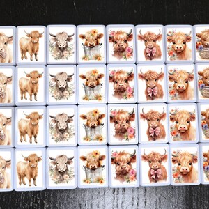 May include: Twenty-four white square tiles with images of Highland cows. Each tile features a different image of a cow with flowers, a bucket, or a basket. The cows are brown and white with pink noses.