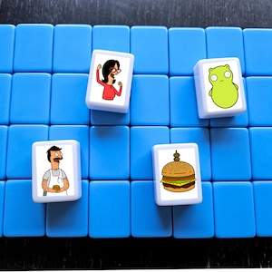 K&ouml;nnte beinhalten: Ein blaues Spielbrett mit vier wei&szlig;en Spielsteinen. Die Spielsteine zeigen Comicbilder eines Burgers, einer Frau mit Brille, eines Mannes mit Schnurrbart und eines gr&uuml;nen Wesens.