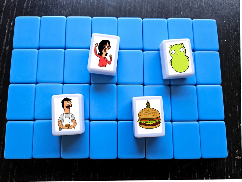 K&ouml;nnte beinhalten: Ein blaues Spielbrett mit vier wei&szlig;en Spielsteinen. Die Spielsteine zeigen Comicbilder eines Burgers, einer Frau mit Brille, eines Mannes mit Schnurrbart und eines gr&uuml;nen Wesens.