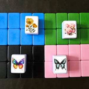 May include: A collection of colorful Mahjong tiles. The tiles are arranged in a grid pattern, with each tile featuring a different floral or butterfly design. The background is a dark surface.