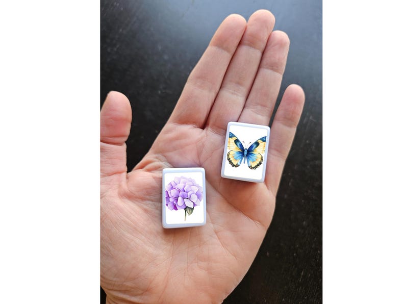 May include: Two small, white, rectangular decorative tiles. One tile features a watercolor image of a purple hydrangea flower. The other tile displays a watercolor butterfly with yellow and blue wings. The tiles are held in a hand.