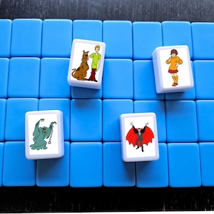 May include: A blue game board with four white tiles. Each tile features a different character from the Scooby-Doo cartoon. The characters are Scooby-Doo, Shaggy, Daphne, and a vampire.