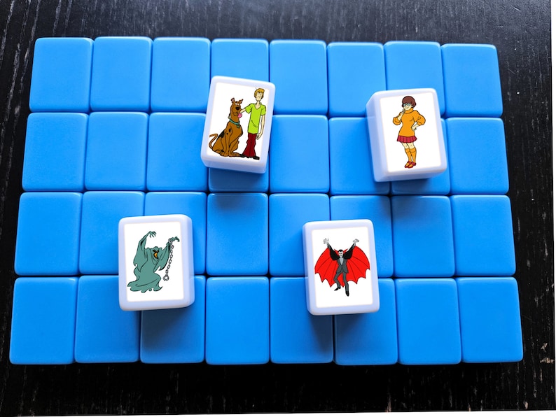 May include: A blue game board with four white tiles. Each tile features a different character from the Scooby-Doo cartoon. The characters are Scooby-Doo, Shaggy, Daphne, and a vampire.