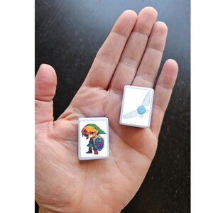 May include: Two white square tiles with images of characters from the Legend of Zelda video game series. One tile features Link, the main character, in green and blue clothing. The other tile features a blue fairy with wings.