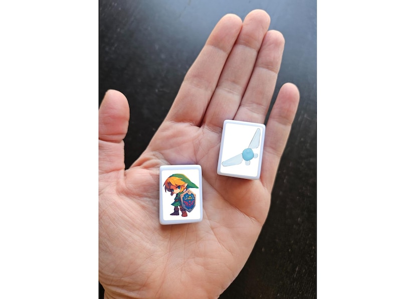 May include: Two white square tiles with images of characters from the Legend of Zelda video game series. One tile features Link, the main character, in green and blue clothing. The other tile features a blue fairy with wings.