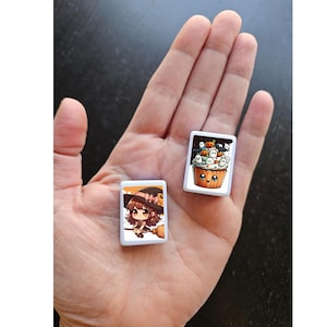 May include: Two white square tiles with Halloween-themed illustrations. One tile features a cartoon witch with a broom and a black hat. The other tile features a cartoon cupcake with a smiling face and a variety of Halloween decorations.