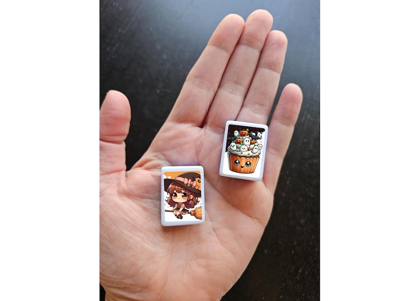 May include: Two white square tiles with Halloween-themed illustrations. One tile features a cartoon witch with a broom and a black hat. The other tile features a cartoon cupcake with a smiling face and a variety of Halloween decorations.