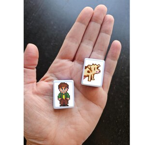 May include: Two small, rectangular pixel art magnets are held in a hand. One features a character in a green shirt and brown pants. The other shows a brown and white cow. The magnets have white borders.