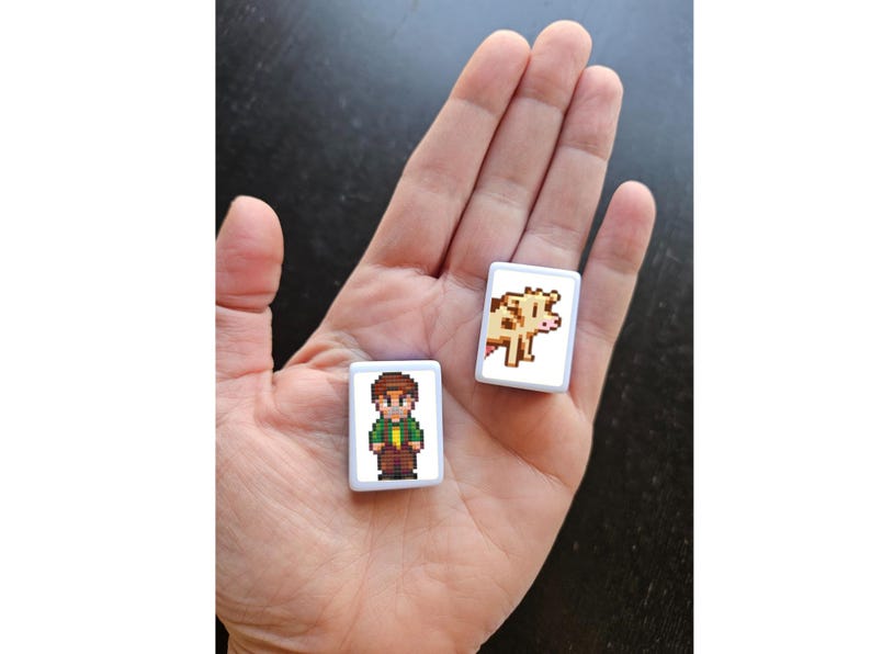 May include: Two small, rectangular pixel art magnets are held in a hand. One features a character in a green shirt and brown pants. The other shows a brown and white cow. The magnets have white borders.