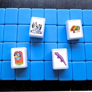 K&ouml;nnte beinhalten: Ein blaues Spielbrett mit vier wei&szlig;en Spielsteinen. Jeder Stein hat ein anderes Cartoon-Bild: zwei Hunde, ein Eidechse und ein Xylophon.
