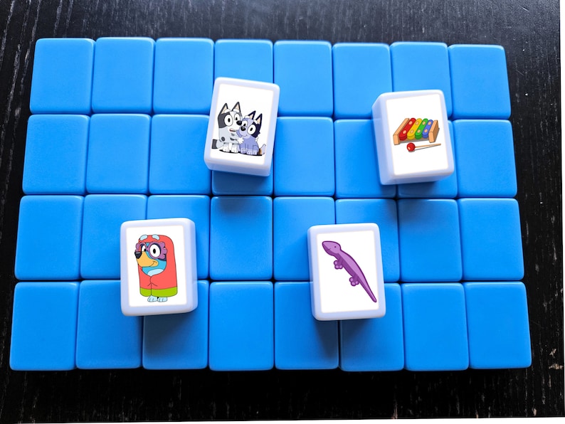 K&ouml;nnte beinhalten: Ein blaues Spielbrett mit vier wei&szlig;en Spielsteinen. Jeder Stein hat ein anderes Cartoon-Bild: zwei Hunde, ein Eidechse und ein Xylophon.