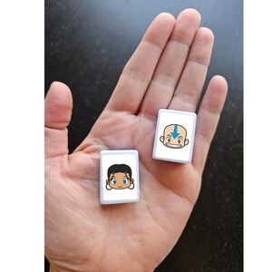 May include: Two small, white rectangular tiles with cartoon character faces are held in a hand. One tile features a bald character with a blue arrow on the forehead. The other tile has a character with black hair and blue eyes. The tiles are likely game pieces.