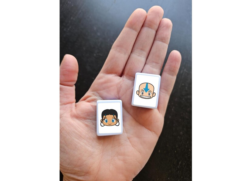 May include: Two small, white rectangular tiles with cartoon character faces are held in a hand. One tile features a bald character with a blue arrow on the forehead. The other tile has a character with black hair and blue eyes. The tiles are likely game pieces.