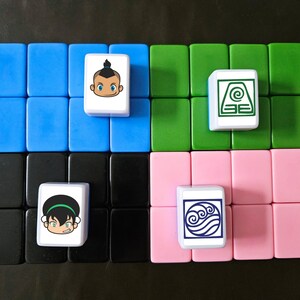 May include: A set of colorful tiles arranged in a grid, with four tiles featuring cartoon character illustrations. The tiles are blue, green, pink, and black, with white tiles displaying character art. The background is a dark surface.