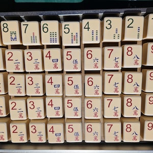 May include: A collection of off-white Mahjong tiles arranged in a metal tray. The tiles feature various numbers and Chinese characters in red and green. The game pieces are ready for play.
