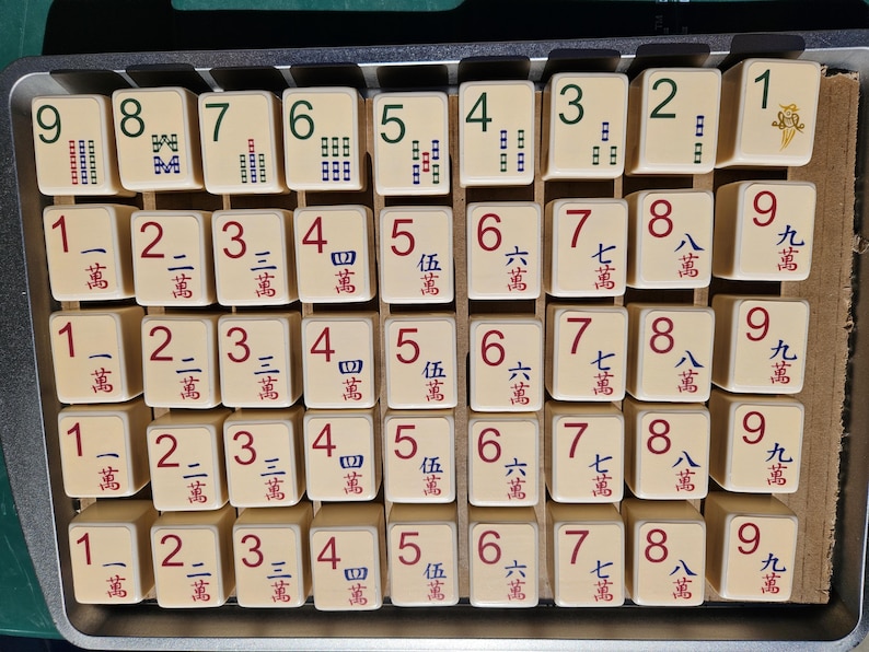 May include: A collection of off-white Mahjong tiles arranged in a metal tray. The tiles feature various numbers and Chinese characters in red and green. The game pieces are ready for play.