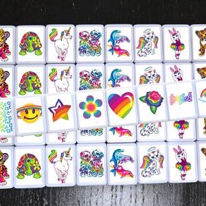 Op de afbeelding: Een set van 25 kleurrijke tegels met verschillende afbeeldingen, waaronder een regenboog hart, een eenhoorn, een bloem, een smiley, een ster, een dolfijn, een kat, een konijn, een tijger, een schildpad en een luipaard. De tegels zijn wit met een zwarte rand.