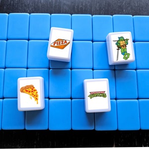 May include: A blue game board with four white tiles. The tiles have images of pizza, a Teenage Mutant Ninja Turtle, and the words "Teenage Mutant Ninja Turtles".