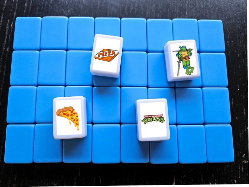 May include: A blue game board with four white tiles. The tiles have images of pizza, a Teenage Mutant Ninja Turtle, and the words "Teenage Mutant Ninja Turtles".
