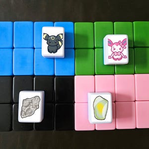 Puede incluir: Un conjunto de cuatro azulejos cuadrados con diferentes im&aacute;genes. Los azulejos est&aacute;n dispuestos en una cuadr&iacute;cula de 2x2. El azulejo superior izquierdo es azul y presenta una criatura de dibujos animados negra y amarilla. El azulejo superior derecho es verde y presenta una criatura de dibujos animados rosa con un lazo. El azulejo inferior izquierdo es negro y presenta una roca gris. El azulejo inferior derecho es rosa y presenta una roca amarilla y verde.