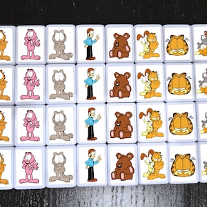 May include: A set of 24 white square tiles with colorful cartoon images of Garfield the cat. The tiles feature Garfield in various poses, including sitting, standing, and holding a plate of lasagna. There are also tiles with Garfield's friends, Odie the dog and Nermal the cat.