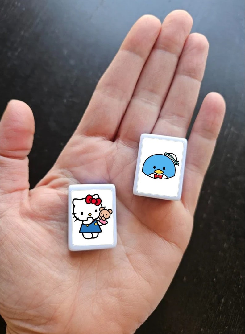 May include: Two white square tiles with cartoon characters. The first tile features Hello Kitty holding a small brown bear. The second tile features a blue penguin wearing a sailor hat and a red bow tie.