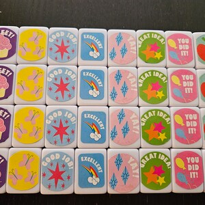 May include: A collection of white tiles with colorful illustrations and text. Designs include cupcakes, butterflies, stars, rainbows, and phrases like "SWEET!", "GOOD JOB!", "EXCELLENT", "GREAT IDEA!", and "YOU DID IT!"