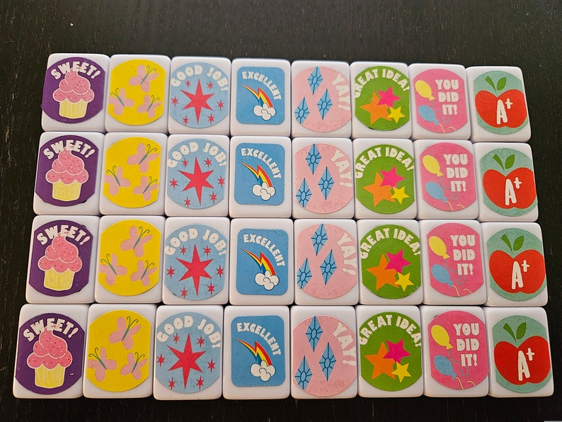 May include: A collection of white tiles with colorful illustrations and text. Designs include cupcakes, butterflies, stars, rainbows, and phrases like "SWEET!", "GOOD JOB!", "EXCELLENT", "GREAT IDEA!", and "YOU DID IT!"