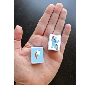 May include: Two white square tiles with blue backgrounds and images of a cartoon pony with a rainbow tail and mane. One tile shows the pony with wings spread, the other tile shows a rainbow with a cloud.