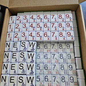 May include: A complete Mahjong set with white tiles, featuring Chinese characters and numbers. The tiles are arranged in a cardboard box, showcasing various suits and directions, including "NESW" and numbers 1-9.