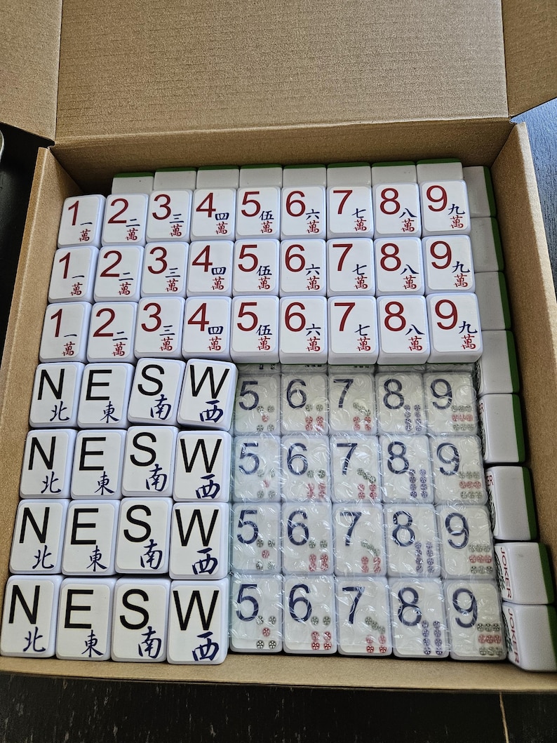 May include: A complete Mahjong set with white tiles, featuring Chinese characters and numbers. The tiles are arranged in a cardboard box, showcasing various suits and directions, including "NESW" and numbers 1-9.