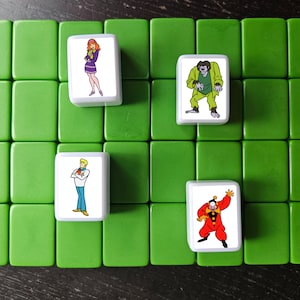 May include: A set of five white tiles with cartoon images of characters from the Scooby Doo franchise. The tiles are on a green surface. The characters include Scooby Doo, Shaggy, Daphne, Fred, and a monster.