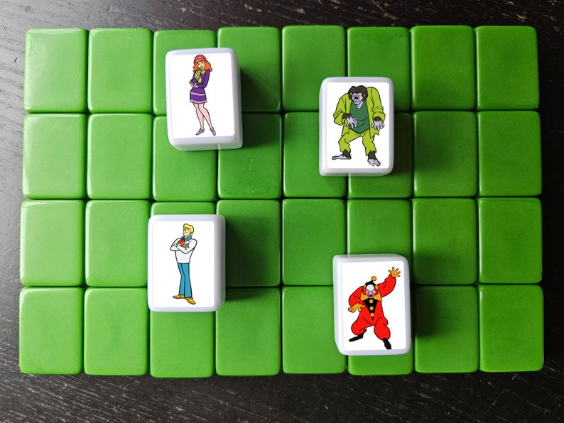 May include: A set of five white tiles with cartoon images of characters from the Scooby Doo franchise. The tiles are on a green surface. The characters include Scooby Doo, Shaggy, Daphne, Fred, and a monster.