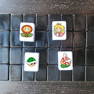 May include: A set of white tiles with images of characters from the Super Mario Bros. video game series. The tiles are arranged on a black background. The characters include a flower, a turtle, a princess, and a plant.