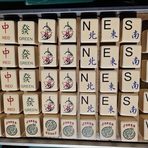 May include: A collection of beige Mahjong tiles with various symbols and characters in red, green, and blue. The tiles are arranged in a metal tray, featuring the words "WHITE", "RED", "GREEN", "JOKER", and directional symbols.