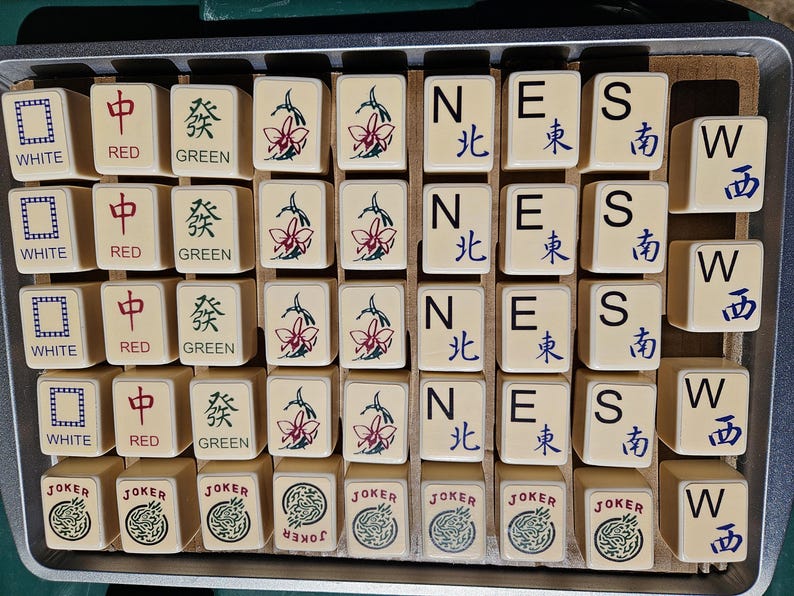 May include: A collection of beige Mahjong tiles with various symbols and characters in red, green, and blue. The tiles are arranged in a metal tray, featuring the words "WHITE", "RED", "GREEN", "JOKER", and directional symbols.