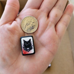 May include: A black and white tile with a black cat wearing a flower crown and a skull. The tile is surrounded by a red and white watercolor background.