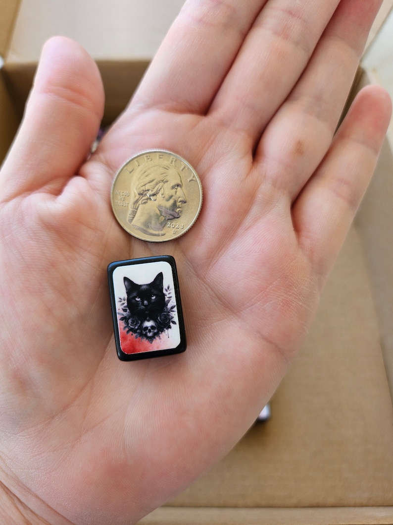 May include: A black and white tile with a black cat wearing a flower crown and a skull. The tile is surrounded by a red and white watercolor background.