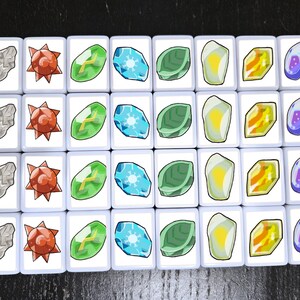Puede incluir: Veinte fichas blancas con diferentes im&aacute;genes de colores. Las im&aacute;genes incluyen una roca gris, una estrella roja, una hoja verde, un copo de nieve azul, un sol amarillo, un diamante naranja y un remolino morado.
