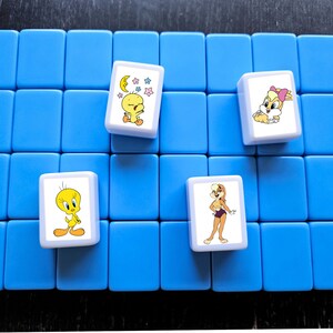 Może przedstawiać: Zestaw niebieskich kafelk&oacute;w do gry z czterema kafelkami z postaciami z kresk&oacute;wek. Postacie to m.in. Tweety, Kr&oacute;lik Lola i inne postacie z Looney Tunes. Płytki ułożone są na niebieskiej powierzchni.