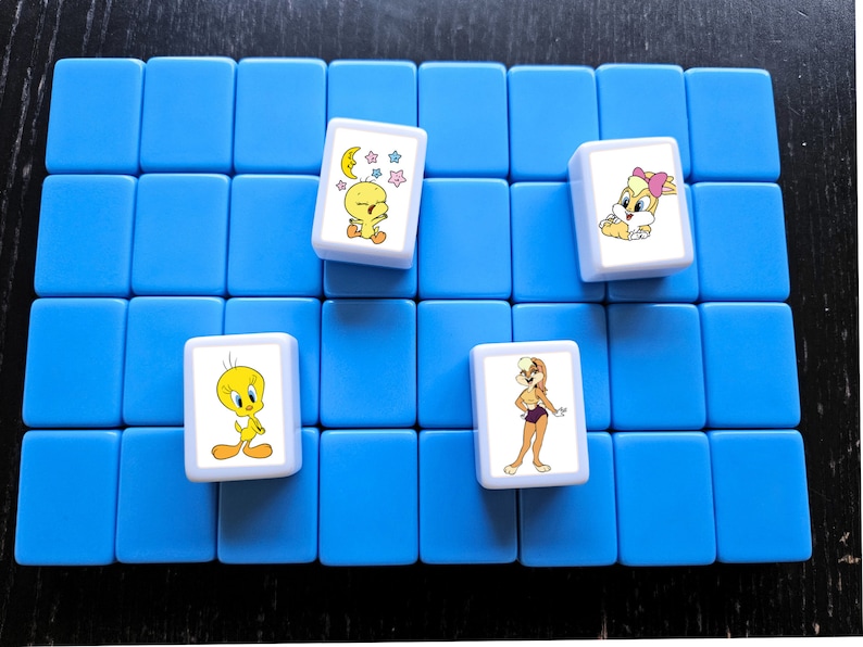 Może przedstawiać: Zestaw niebieskich kafelk&oacute;w do gry z czterema kafelkami z postaciami z kresk&oacute;wek. Postacie to m.in. Tweety, Kr&oacute;lik Lola i inne postacie z Looney Tunes. Płytki ułożone są na niebieskiej powierzchni.