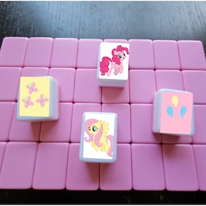 May include: Four white square tiles with different images on them. The tiles are on a pink surface. The images are of a pink pony, a yellow pony, three pink butterflies on a yellow background, and a pink pony with two balloons.