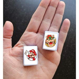 May include: Two white square tiles with images of Mario and Bowser from the Super Mario Bros. video game series. The tiles are on a hand.