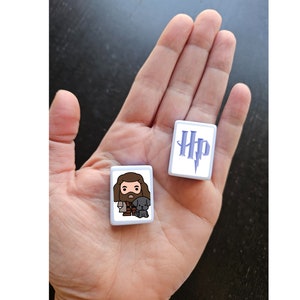 May include: Two white square tiles with images from the Harry Potter universe. One tile features the Harry Potter logo with a lightning bolt in purple. The other tile features a cartoon image of  Rubeus Hagrid with his pet dog Fang.